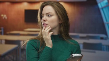 Binada elleri çenesinde akıllı telefon tutan yeşil kazak giyen genç beyaz bir kadın..