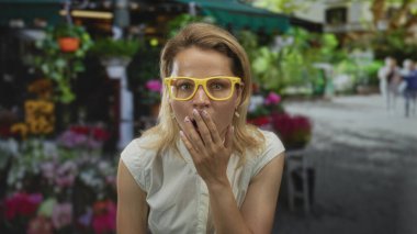 Sarı gözlüklü kadın renkli çiçeklerin yanında kaldırımda dururken ağzını eliyle kapatıyor..