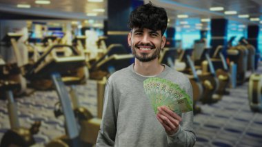 Spor salonunda spor aletleriyle Rus rublesi taşıyan bir adam, kapalı bir spor salonunda güvenle gülümsüyor..