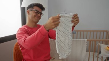 Yatak odasında kucağında bir bebek elbisesi tutan genç bir adam, sıcak aydınlatılmış bir çocuk yuvasında ebeveynlik için mutluluk ve beklenti gösteriyor..