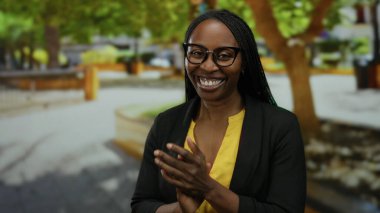 Açık hava parkında kıvırcık saçlı gülümseyen, gözlüklü ve sarı tişörtlü, canlı yeşil bir çevrede ağaçlarla çevrili, neşe ve rahatlama gösteren bir kadın..