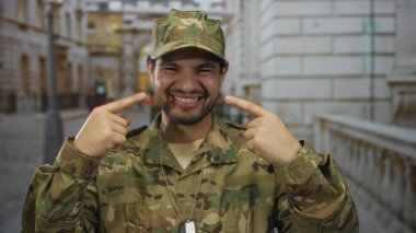 Kamuflaj üniformalı bir adam, şehir sokaklarında güler yüzlü ve yanakları işaret eder..