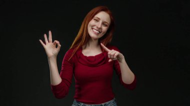 Siyah arka planda gülümseyen kızıl saçlı iyi niyetli bir kadın kırmızı kazak ve kot pantolon giyerken kendine güveni ve pozitifliği sembolize eder..