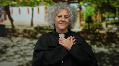 Gri saçlı kadın rahip, açık hava parkında elleri göğsünde, huzurlu bir şekilde gülümsüyor. İnancı ve huzuru temsil ediyor..