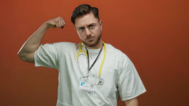 Sakallı genç doktor, beyaz önlük ve steteskopla kırmızı duvara yaslanıyor. Sağlık sektöründe güç ve kararlılık gösteriyor..