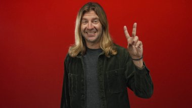 Kırmızı stüdyoda iki eliyle barış işareti yapan adam geniş bir sırıtış, uzun saçlar ve rahat duruş; neşe.