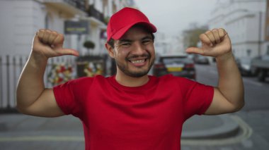 İşlek caddede güpegündüz gülümserken kırmızı şapkanın altında parmaklarını göğsüne gösteren İspanyol bir adam..