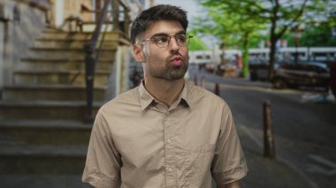 Yuvarlak gözlüklü, bej gömlekli genç İspanyol adam işlek şehir caddesinde dudaklarını büzüyor..