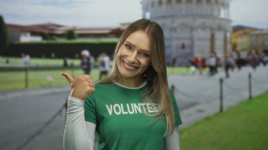 Sarışın kadın gönüllü olarak sidik kulesinin önünde gülümsüyor ve başparmağıyla işaret ediyor. Arka planda insanlar var. Neşe ve toplum ruhunu ünlü bir İtalyan açık hava ortamında aktarıyor..