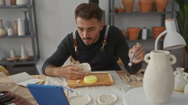 Kil kasesini çıplak elle şekillendiren adam, sanat atölyesinde çömlek raflarıyla ahşap masa üzerinde, yaratıcılığa odaklanıyor..
