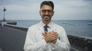 Laboratuvar önlüklü genç İspanyol bilim adamı her iki elini göğsünün üzerinde tutuyor ve gün ışığında deniz kenarında bir sokakta gülümsüyor; minnettarlık.