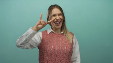 Beyaz tişörtün üzerine pembe kazak giyen gülümseyen sarışın kadın yeşil bir duvar arkasında duruyor, kendine güveni ve yaklaşımı var.