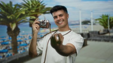 Sahilde gülümseyen yakışıklı bir adam elinde kamera palmiyeleri, sahilde tatil havası, rahatlamış genç, neşeli bir yetişkin ifadesi ile eğleniyor..