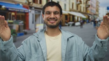 Açık mavi ceketli adam, güneş ışığında hareketli şehir caddesinde, mağaza önü tentelerinin altında kollarını geniş tutuyor..