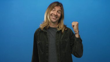 Sarışın, uzun saçlı, gülümseyen bir adam mavi stüdyoda yumruğunu sıkıyor. Zafer kutlamaları için iyimserliği artırıyor..