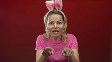 Pembe gömlekli ve tavşan kulaklı genç sarışın kadın kırmızı stüdyoyu işaret ediyor..