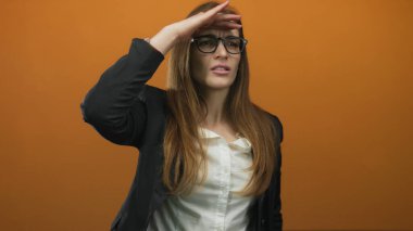 Stüdyoda turuncu duvara bakarken gözlük takan genç bir kadın gözlerini eliyle gölgeliyor..