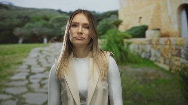 Parkta taş yolu ve yeşilliği olan bir kadın, kırsal manzaranın ortasında tarafsız ifadeler sergiliyor, basitliği ve doğal çevreyi vurguluyor..