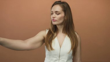 Beyaz üst puanlı kadın stüdyoda çıplak koldan yana parmağıyla turuncu arka planda; kararlılık özgüveni berraklığı yönü.