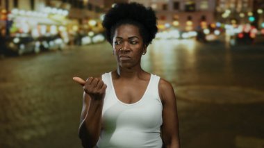 Beyaz atletli bir kadın gecenin bir yarısı hareketli bir caddede duruyor, sokak lambalarının altında düşünceli bir ifadeyle kenarı işaret ediyor..