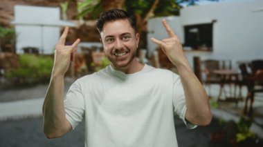 Sakallı genç adam terasta kahveyle gülümsüyor şehir sokaklarında, rahat bir restoran ortamında şehir yaşamını sergiliyor..