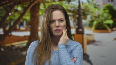 Mavi kazak giyen kadın yeşil bir park yolunda ürkerken yanağa dokunuyor; diş ağrısı.