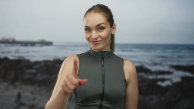 Kolsuz atletik bluzlu bir kadın sahilde el hareketi yapıyor ve okyanus dalgaları görünüyor. Eğlenceli bir davet..