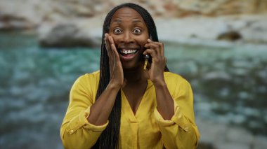 Arka planda deniz kenarı olan akıllı telefondan konuşan bir kadın, kumsalda açık sarı kıyafetli bir telefonla eğleniyor..
