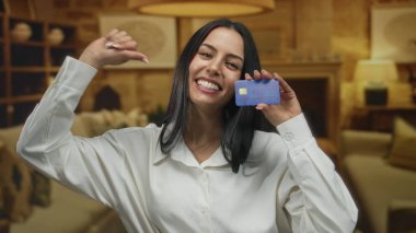 İçerideki İspanyol kadın rahat bir otel ortamında gülümseyip kendini gösterirken aynı zamanda gururla kredi kartı gösteriyor, boş zamanlarını ve seyahatleri vurguluyor..