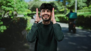 Açık hava parkında gülümseyen genç bir adam, yemyeşil ile çevrili, güneşli bir günün tadını çıkarıyor, neşeli bir jest ile huzurlu ve doğal bir ortamda yakalandı..