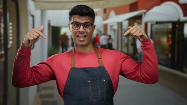 Canlı bir sokak ortamında kırmızı bir kazak ve kot önlük giymiş, kendine güveni ve modern şehir tarzı açık hava tarzıyla gülümseyen genç İspanyol bir adam..