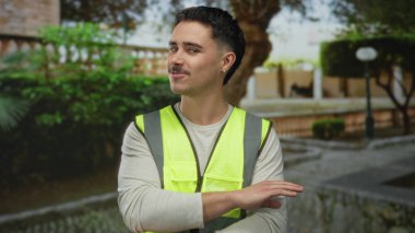 Gündüz vakti yeşil ve tuğla duvarlarla çevrili şehir sokaklarında yansıtıcı yelek giyen genç İspanyol adam güvenlik ve şehir ortamını gösteriyor.