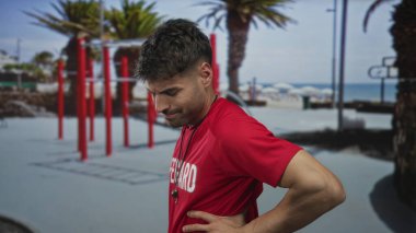 Genç adam, palmiye ağaçları ve kırmızı egzersiz çubuklarının yakınındaki güneşli şehir sokağında belini tutuyor..