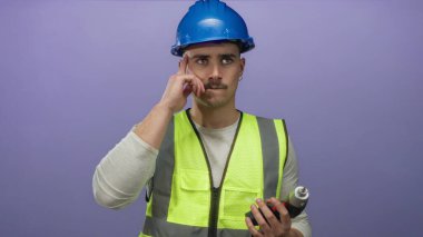 Yansıtıcı yelek ve şapka giymiş genç İspanyol bir adam mor bir arka plan duvarına karşı matkap tutuyor, düşünceli ve odaklanmış görünüyor..