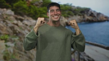 Hispanik adam açık gökyüzü altında rüzgarlı kayalık sahilde yumruk sallar ve yumruklarını sıkar; zafer.