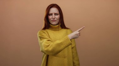 Sarı bir süveteri basit bir kahverengi duvar arka planına doğrultup kafa karışıklığını ifade eden bir kadın, kırmızı saçlarına ve günlük tarzına odaklanmış duygu jestleri sergiliyor.