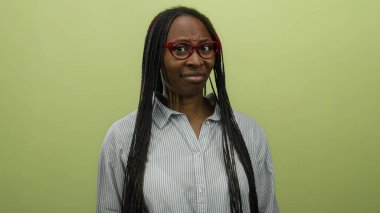 Örgülü ve gözlüklü bir kadın sarı arka plana şüpheyle bakıyor, çizgili bir gömlek giyiyor, merakını iletiyor ve bakışlarını sorguluyor..