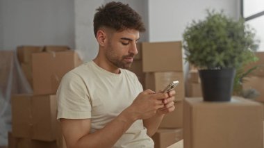 Akıllı telefon ve kaydırmalı ekran taşıyan adam modern binanın içinde istiflenmiş karton kutuların yanında; yeni bir başlangıç umudu.