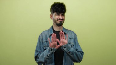 Tereddüt ve belirsizliği vurgulayan, parlak sarı bir arkaplan önünde kot ceket giyen, şüpheci bir ifadeyle isteksizliğini ifade eden genç bir adam..