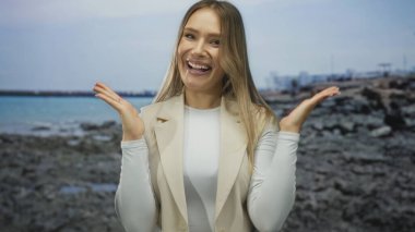 Gün ışığında kayalık bir sahilde neşeli bir şekilde gülümseyen kadın bej bir yelek ve beyaz bir kıyafet giyiyor, berrak bir gökyüzünün altında huzurlu denizin yanında mutluluk ve rahatlığı somutlaştırıyor..