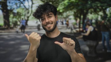 Sakallı ve siyah gömlekli genç adam yeşil park ortamında parmaklarını yana doğru uzatarak neşeli bir ifade ve şehir ortamında rahat bir ruh hali sergiliyor..