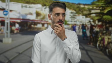 Beyaz gömlekli adam, odaklanmış bakışları ve hafif çizik kaşları ile parlak güneş ışığı altında, sokakta düşünceli bir jest çene dokunur;.