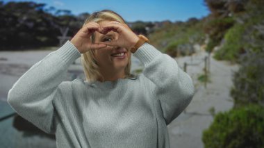 Genç sarışın kadın kalp hareketi yapıyor elleri çerçeveli açık hava parkında parlak güneş ışığı altında göz; aşk bağlantısı.