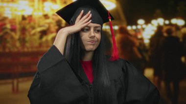 Mezuniyet kepi ve cüppeli İspanyol kadın, arka planda parlak ışıklar ve coşkulu kalabalıkla açık hava fuarında düşünceli görünüyor..