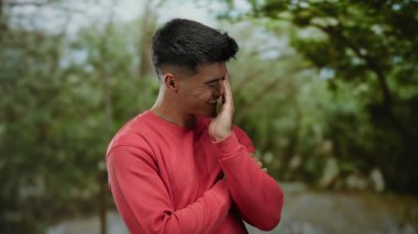 Genç adam canlı park ortamında gülüyor, kırmızı tişört giyiyor, yemyeşil bir zemin ve yumuşak doğal ışığa karşı samimi bir neşe anında yakalandı.