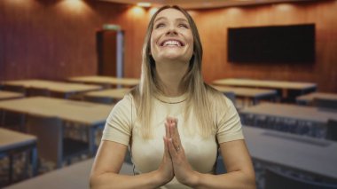 Sınıfta dua ederken sımsıkı gülümseyen bir kadın hafif ışıklandırma ve boş masalarla geliştirilmiş gençlik pozitifliğini ve modern öğrenme ortamını yansıtan masalar..