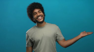 Mavi arka planda gülümseyen kıvırcık saçlı ve günlük giysileri olan bir adam izole edilmiş stüdyoda mutlu bir şekilde poz veriyor ve pozitif bir ifade sergiliyor.