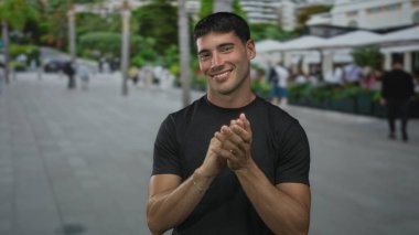 Young hispanic man wearing black shirt clapping hands and smiling on street outdoors; celebration joy.
