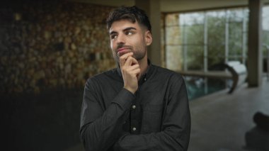 Çene üzerinde eli olan genç İspanyol adam kapalı bir havuz yanında mozaik bir duvar önünde duruyor; sakin yansıma.