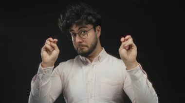 Sakallı ve gözlüklü genç bir adam siyah bir arka plana, bir stüdyo ortamında yüz ifadesine ve jestlere karşı tırnak işareti yapıyor..